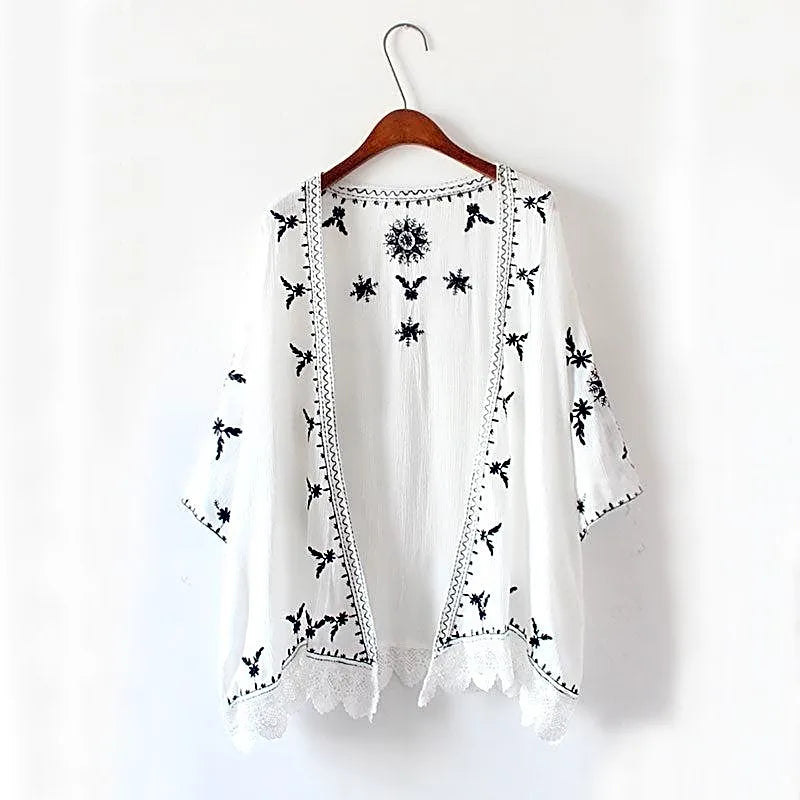 Biała narzutka kimono w stylu boho z czarnym haftem i koronkowym wykończeniem, wisząca na wieszaku, widok z przodu.