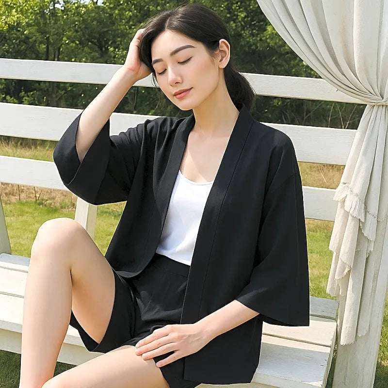 Młoda kobieta w czarnej eleganckiej kurtce kimono z szerokimi rękawami, siedząca na ławce w ogrodzie, idealna jako letnia narzutka.