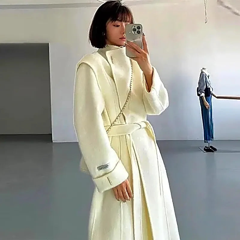 Długi kremowy płaszcz damski w stylu kimono z wysoką stójką i wiązanym paskiem, widok na modelce.