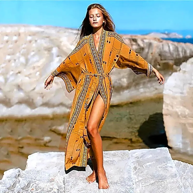 Długie pomarańczowe kimono plażowe boho z etnicznymi geometrycznymi wzorami i paskiem, noszone przez modelkę na tle skalistego wybrzeża.