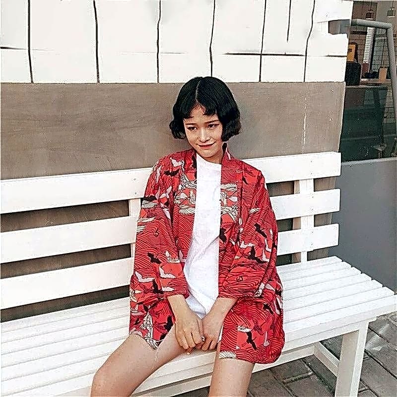 Krótkie czerwone kimono damskie z motywem lecących żurawi, stylizacja casualowa na siedzącej modelce.